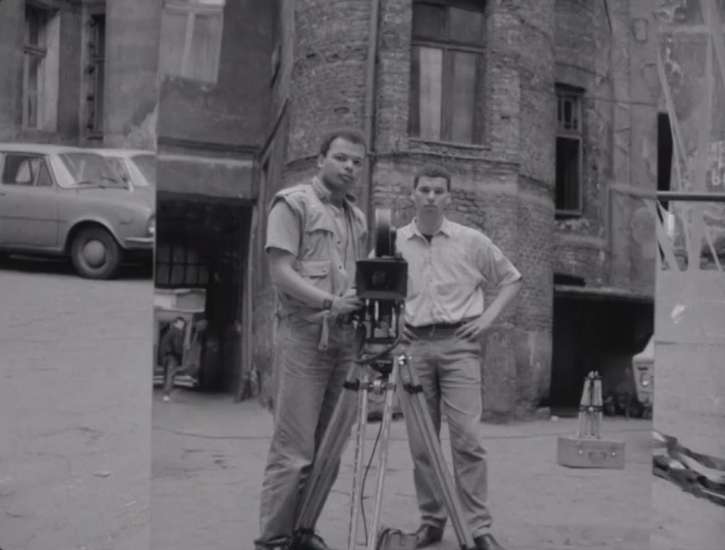 Two men standing next to a film camera on a city street looking at are reflected