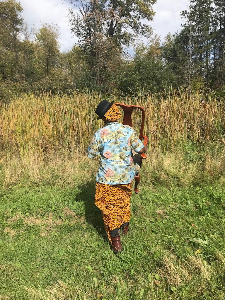 A person with dark skin wearing printed fabrics and a black hat walks on grass away from the camera, carrying a wooden chair towards a patch of tall grasses.