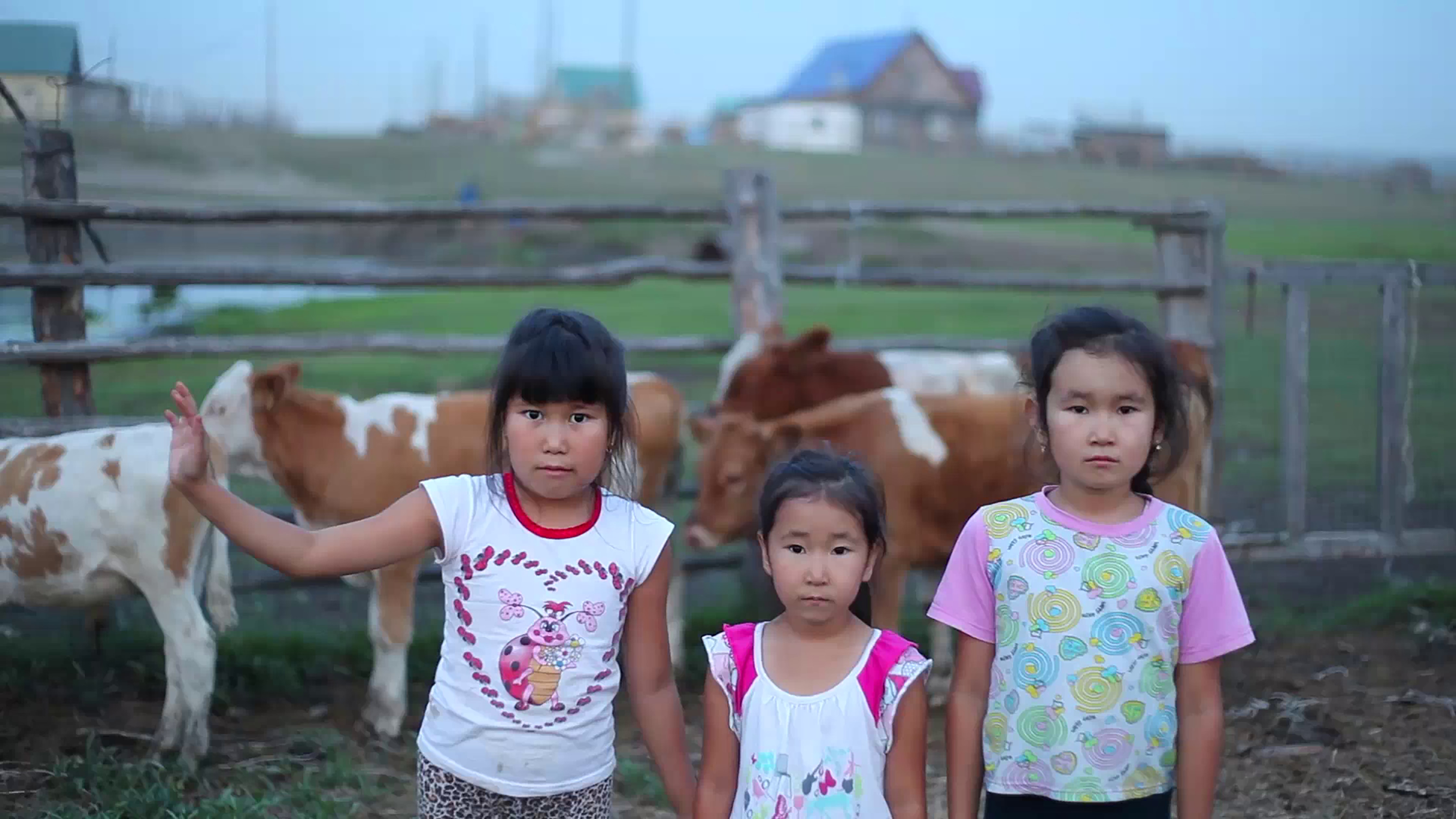 Three children face the camera, standing close together in front of cows and a field with distant houses.