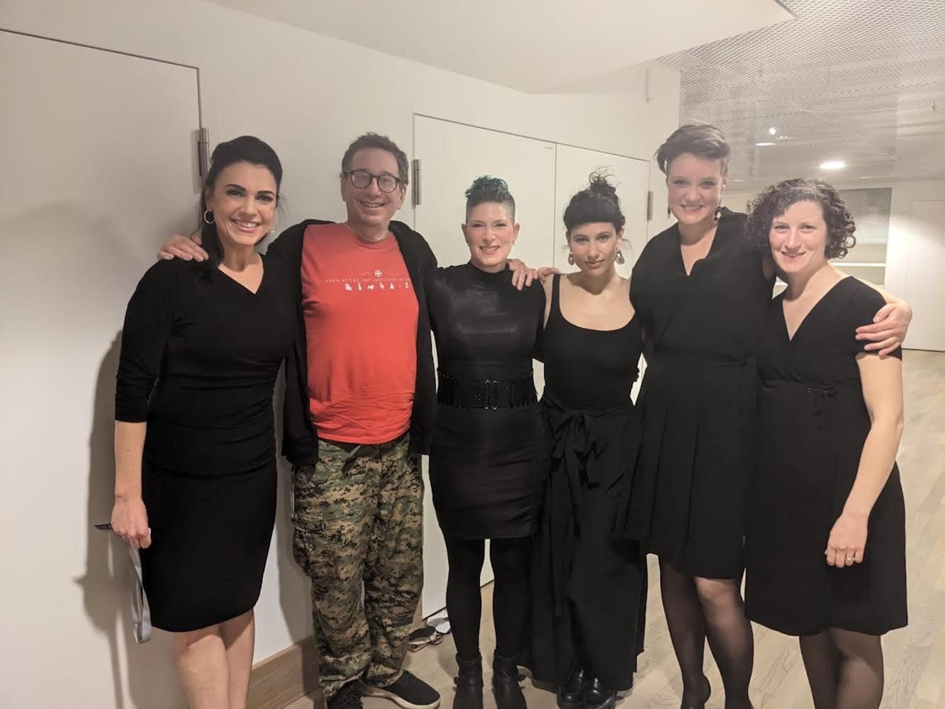 Five women and one man stand in a row arms on each other and they look and smile at the camera. The five women all wear stylish black dresses of varying styles. The man wears glasses, a red tee-shirt, and black hoodie and camo pants. They are all standing in a white room with many white doors and a low shimmering ceiling.