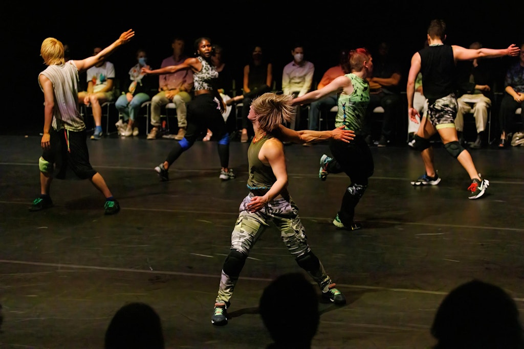 Five dancers in various colors of streetwear, knee pads, and sneakers swinging their right arms behind them. They dance energetically in front of a row of audience members sitting on chairs along the back of the stage.