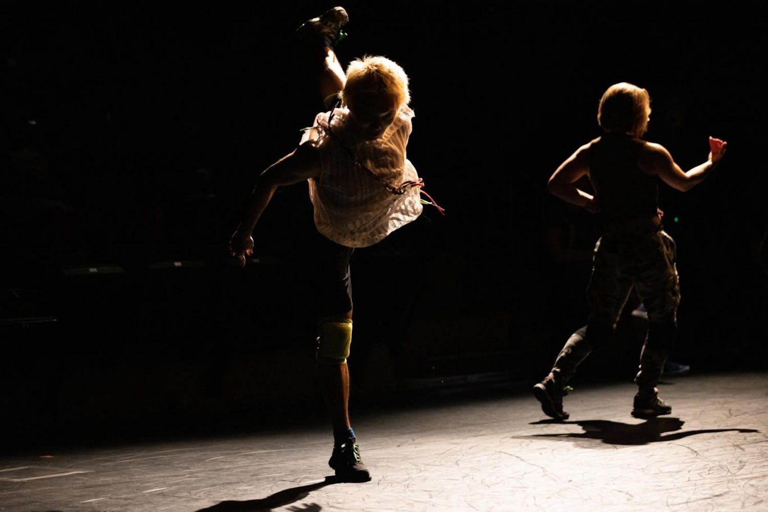 On a darkly lit stage and both in silhouette, one dancer stands on one leg and the other faces away from the camera.