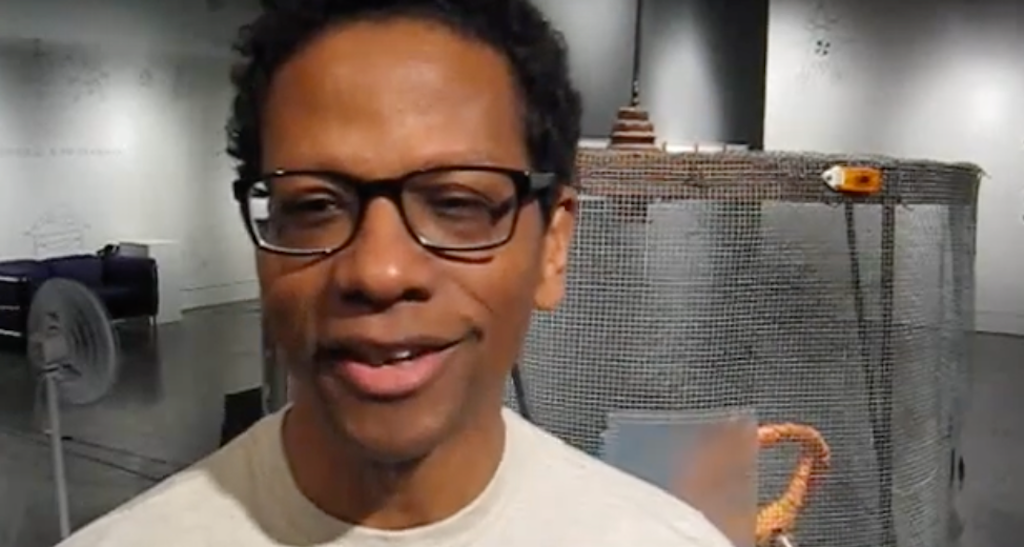 An adult with dark skin and dark rimmed glasses faces the viewer in mid-speak.