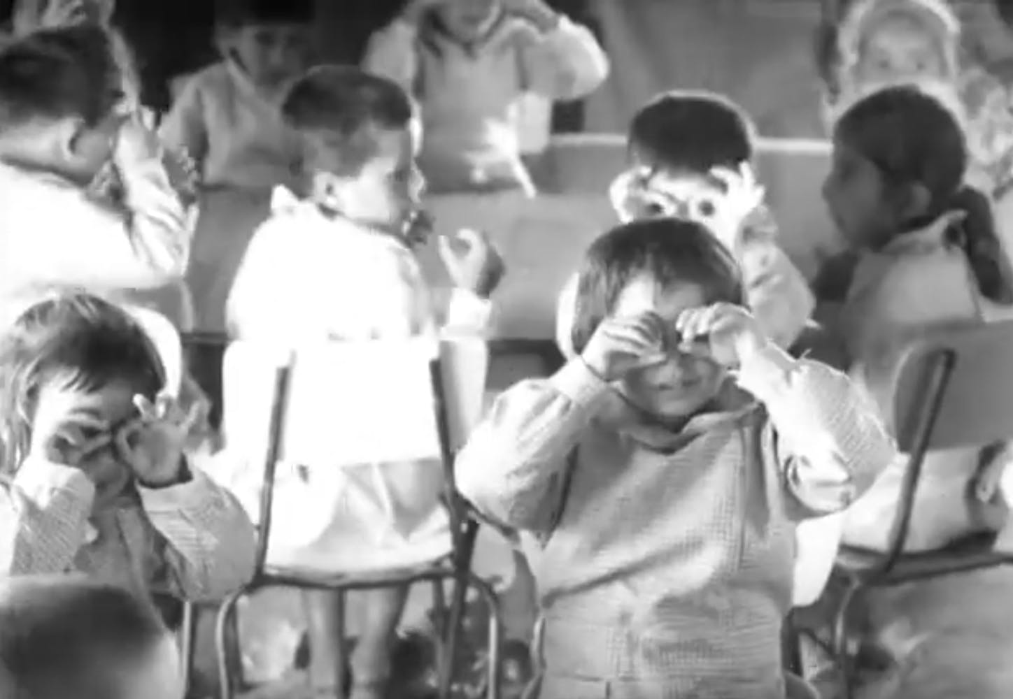 Black and white image of a group of children sitting in classroom desks and holding their hands up to their eyes to mimic glasses.