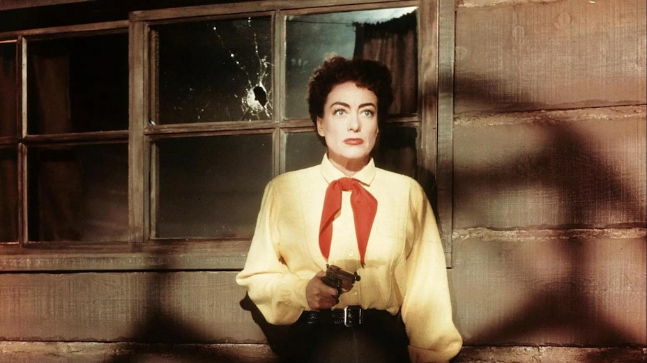 A woman with short dark curly hair wearing a yellow shirt and red neckerchief holds a handgun while standing against a house with a broken window.