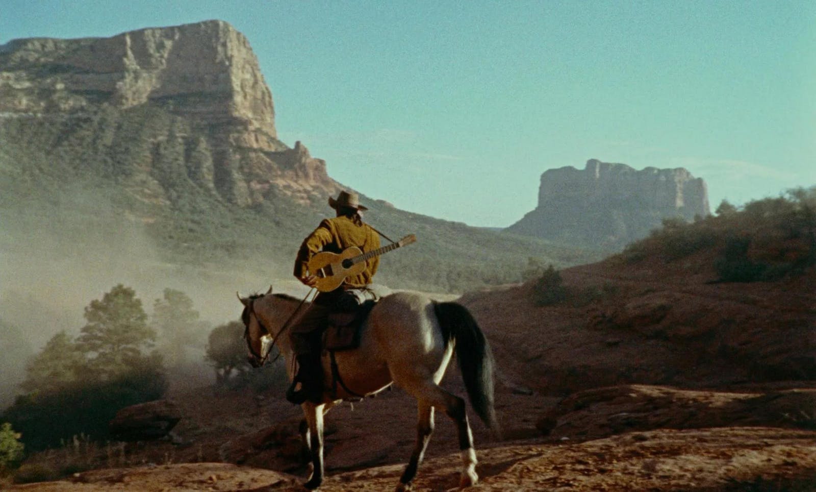 A man with a guitar on his back rides off on horseback among a dusty hazy landscape of mountainous red rock formations on a bright blue sky.