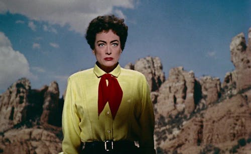 A woman with short curly hair and red lipstick wearing a yellow shirt and red neckerchief has a stern look on her face. She stands in a rocky landscape.