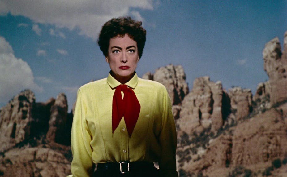 A woman with short curly hair and red lipstick wearing a yellow shirt and red neckerchief has a stern look on her face. She stands in a rocky landscape.