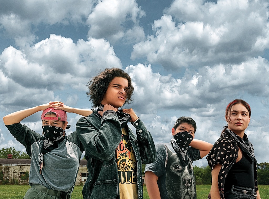 Four teenagers outdoors with cloudy blue sky above them. They wear black bandanas, two wear them over their faces, and black, gray and denim clothing.