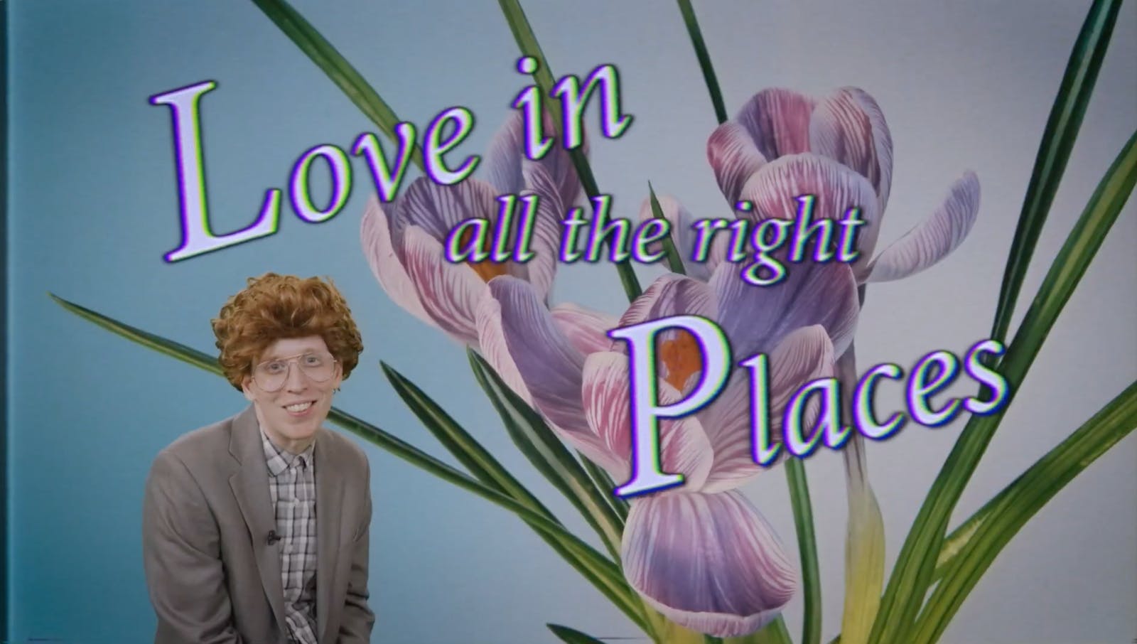 A man in 1970's glasses and a wig smiles at the viewer while sitting in front of an image of flowers witht he text 