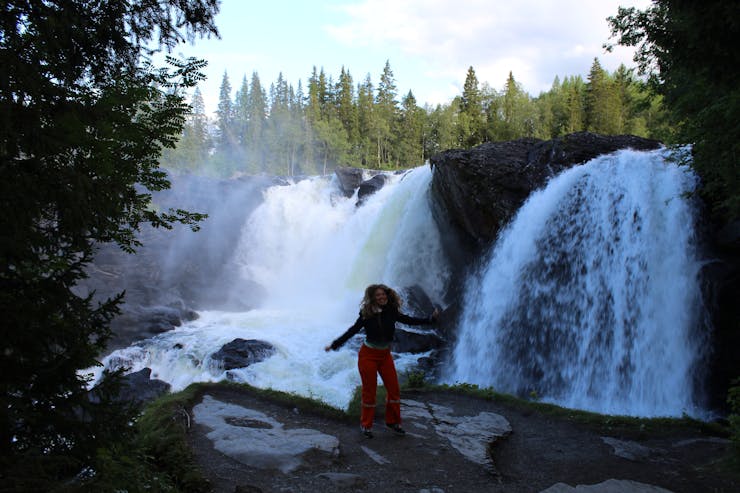 Parker Genné, a woman with long brown curly hair, moving in front of a large Scandinavian waterfall. She wears red pants and a brown jacket.
