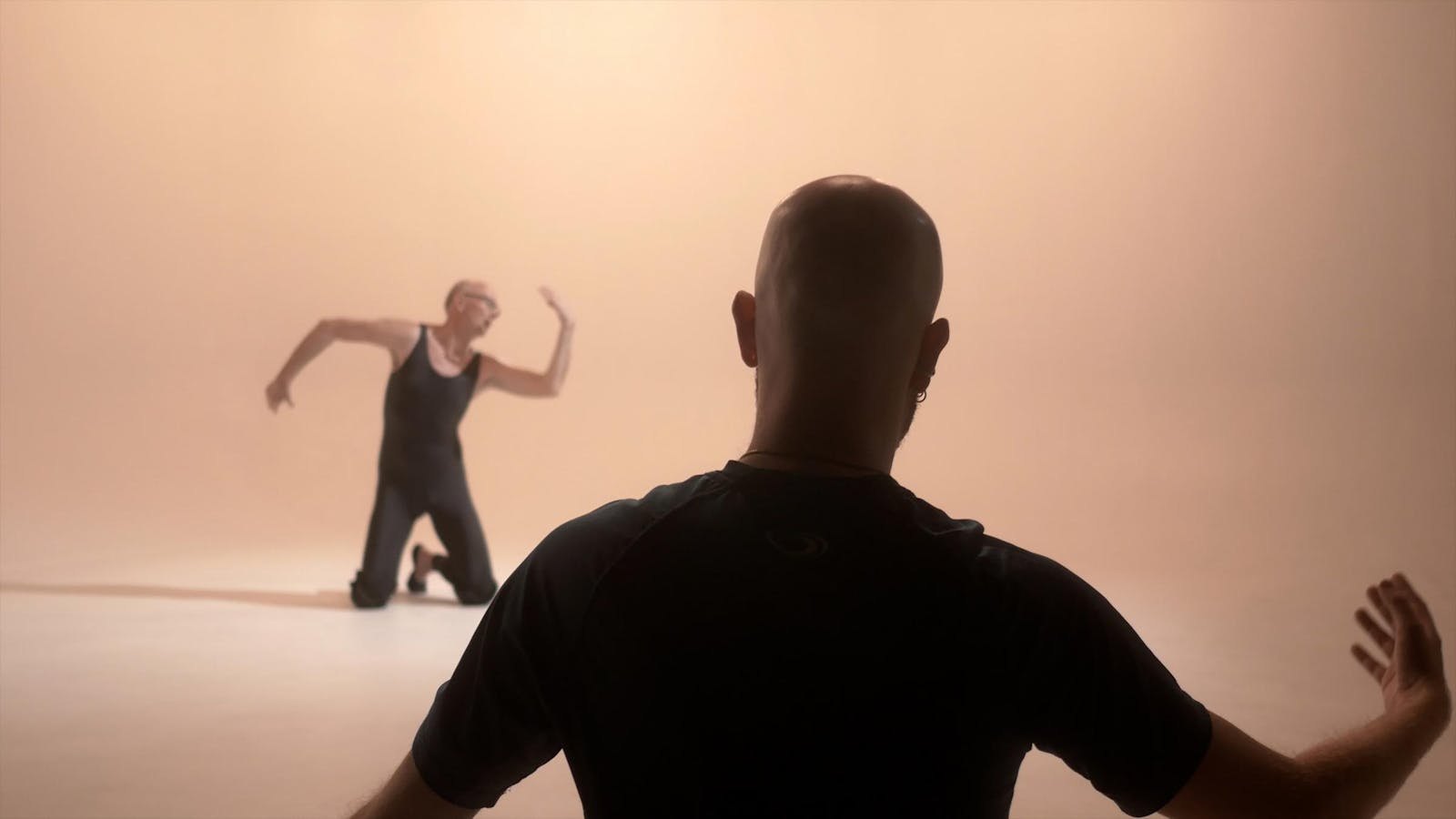 In a soft beige room, an older man in the distance strikes a ballet pose on his knees. A man mimics his pose in the foreground.
