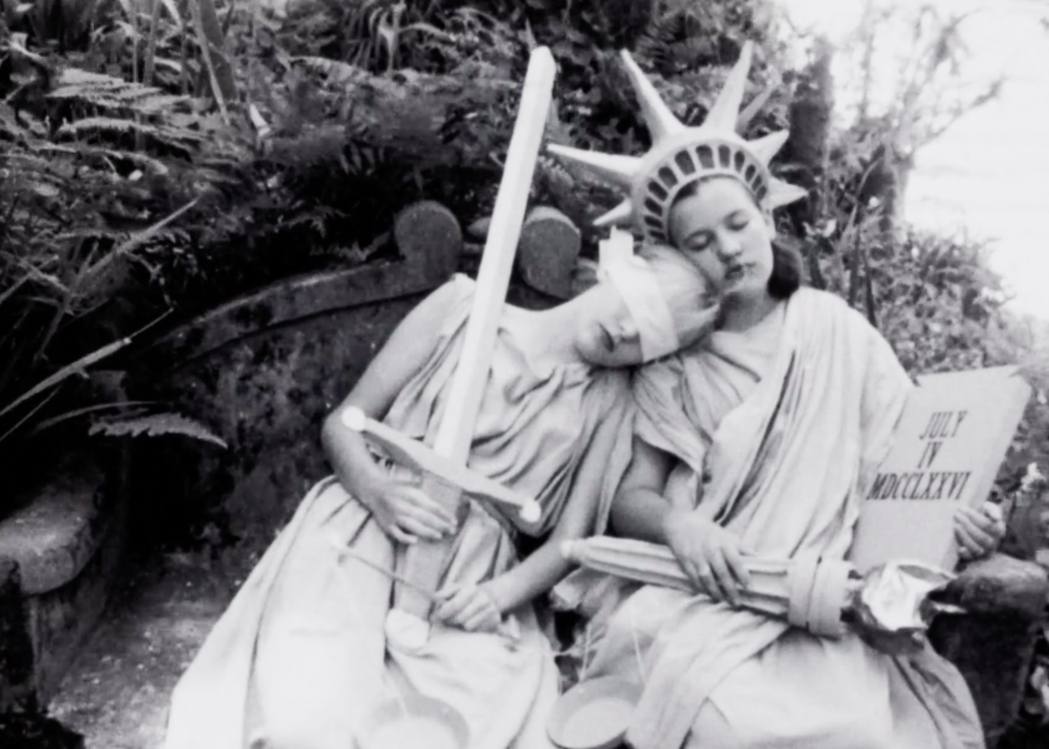 Two women sitting next to each other in togas. One wearing a blindfold holding a sword and scale, the other in the Statue of Liberty attire (crown, torch).