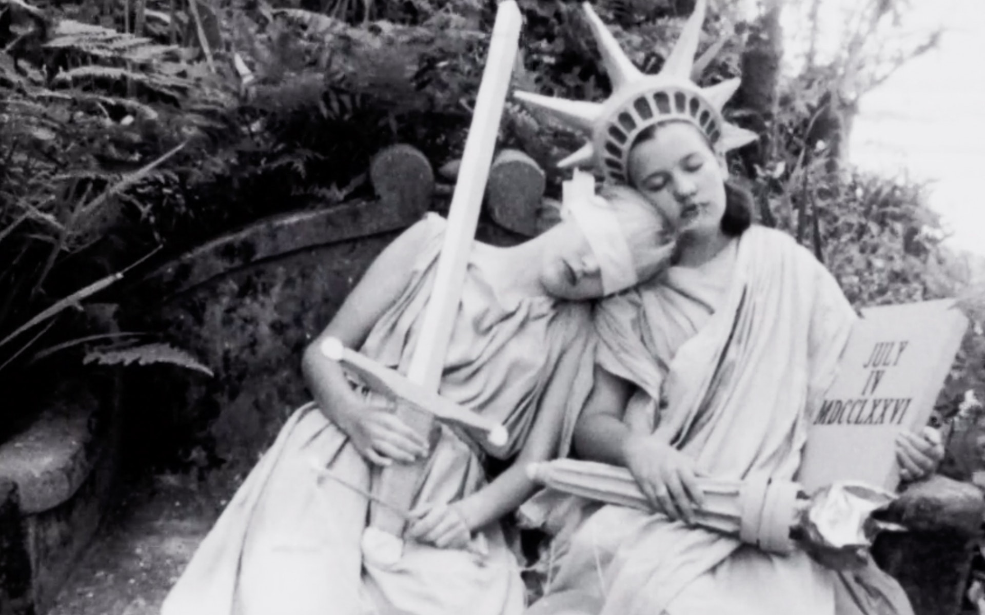 Two women sitting next to each other in togas. One wearing a blindfold holding a sword and scale, the other in the Statue of Liberty attire (crown, torch).