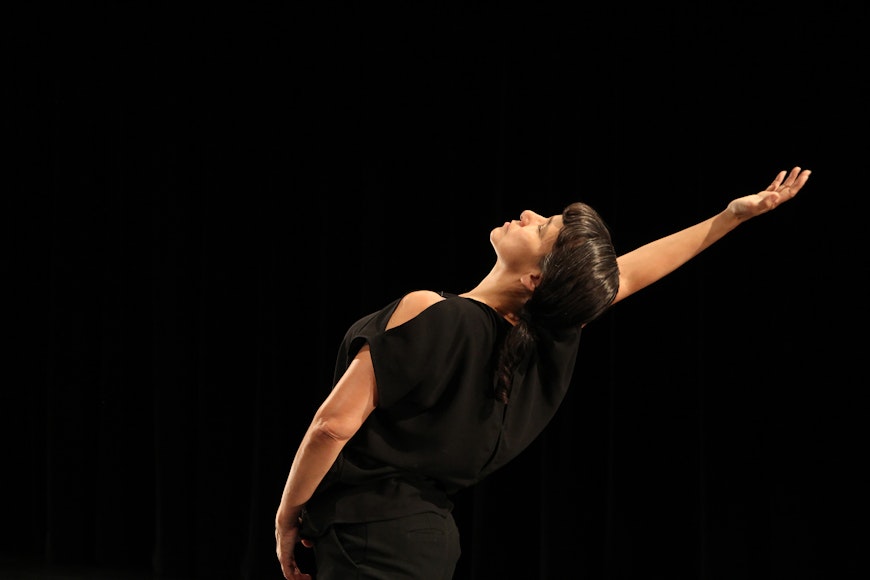 A dancer wearing all black extends her arm backward against a black background.