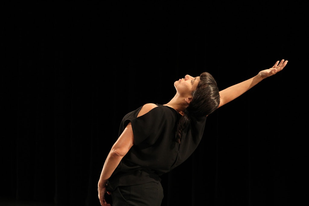 A dancer wearing all black extends her arm backward against a black background.