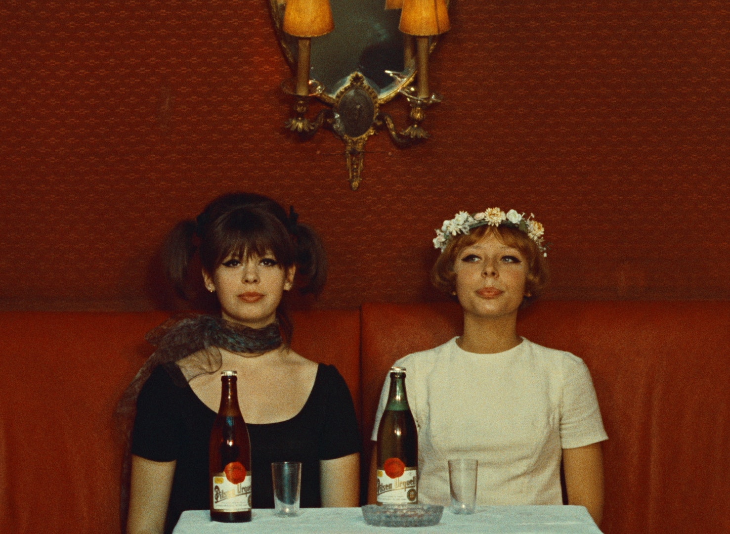 Two adult woman with light skin, one wearing white with flowers in their hair and one wearing black, sit next to one another in a bar booth with two bottles of beer and glasses in front of them.