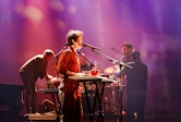 A group of five musicians stand on stage, awash in purple light. A woman in a jumpsuit plays the keyboard and sings in the center of the photo.