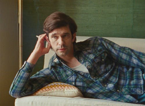 A man with light skin and brown hair lays on a couch in a green room on his side, propped on his elbow and holding a cigarette. He wears a blue plaid shirt.