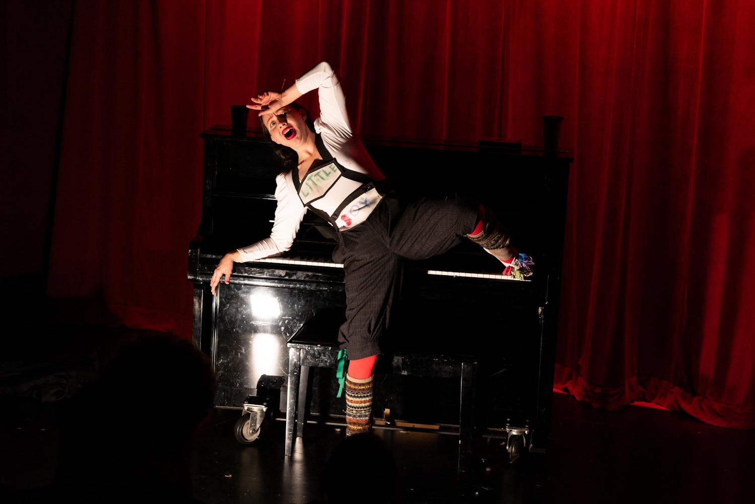 A performer with light skin wearing lederhosen pretends to faint on an upright piano.