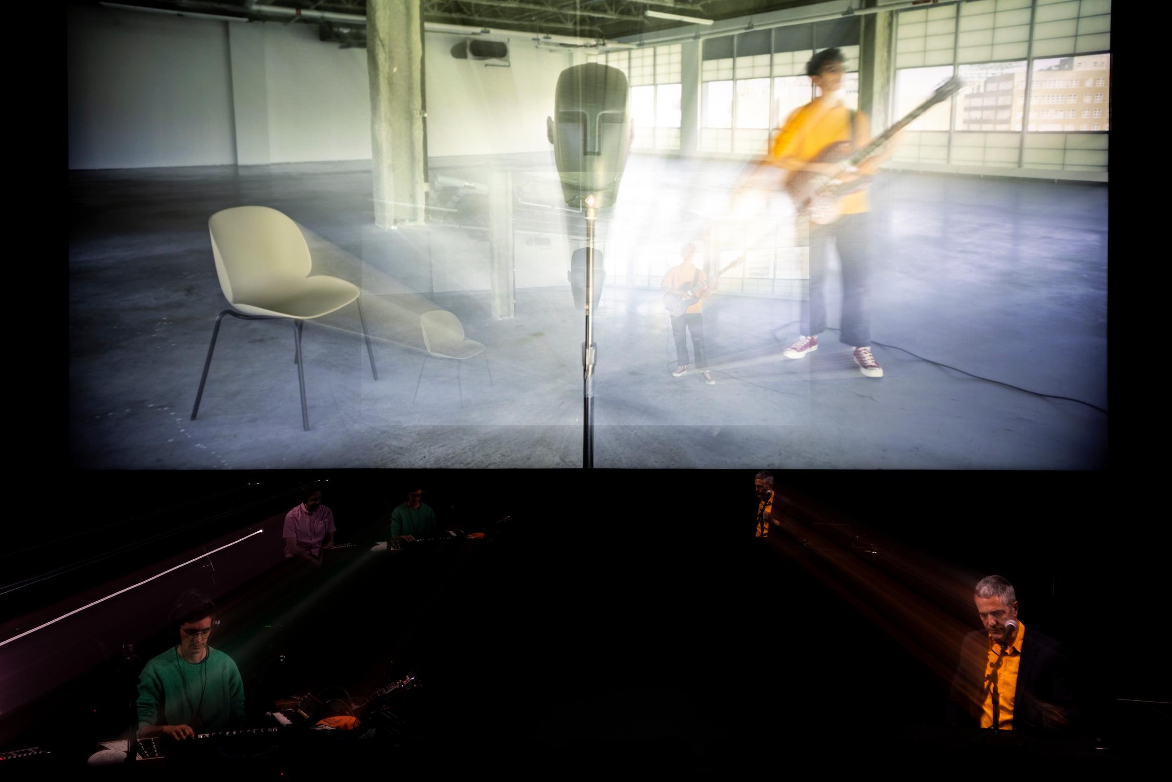 Two people sing and play music on from of a projection of someone playing guitar.