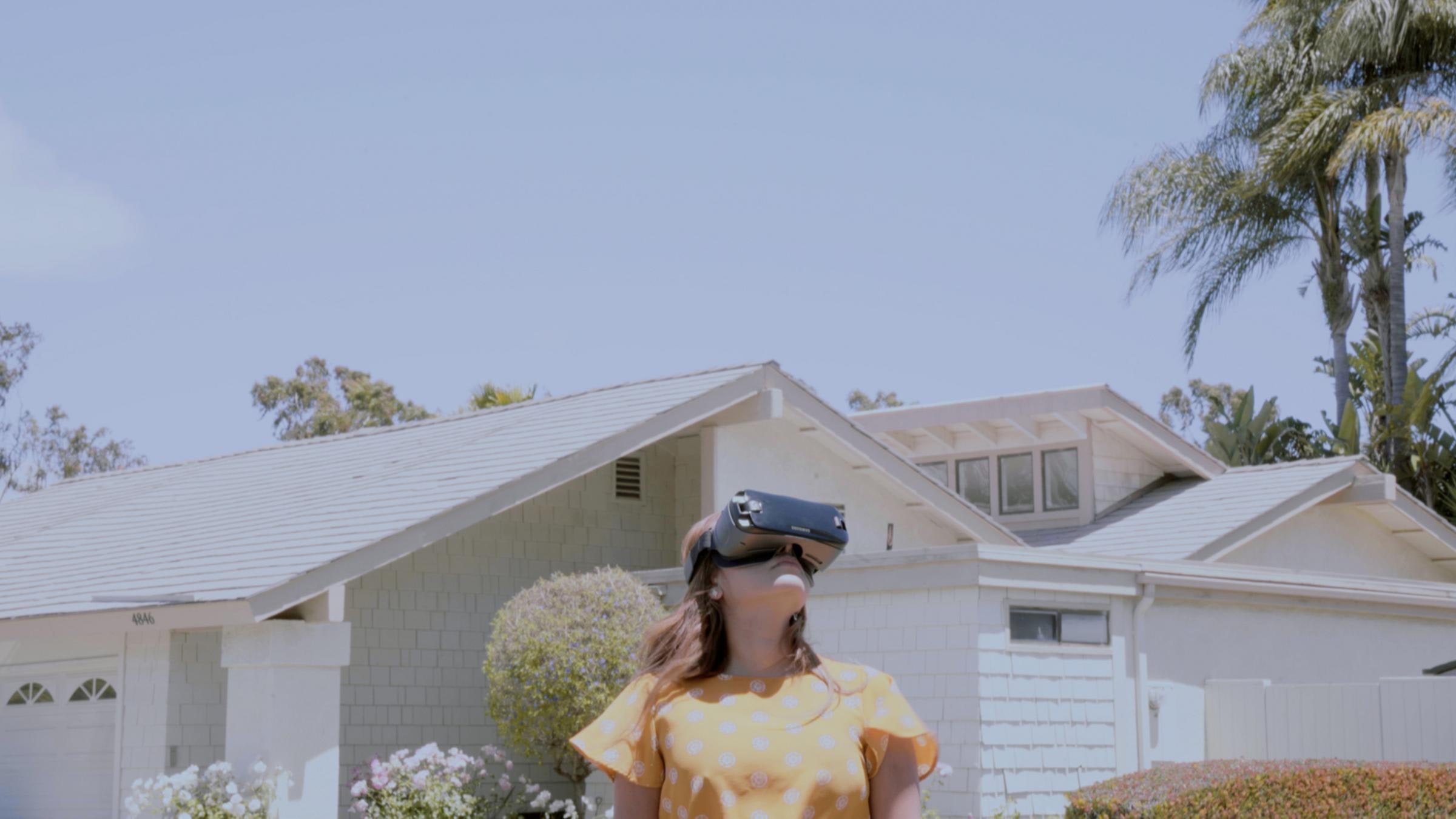 A woman wearing a virtual reality headset and dressed in a yellow polka dot top stands in front of a house and looks upward toward the sky.