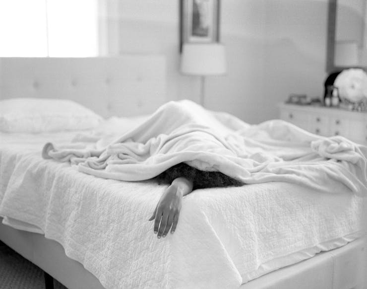 A black and white image. A dark-skinned hand extend out from under a blanket on a bed in a bedroom.