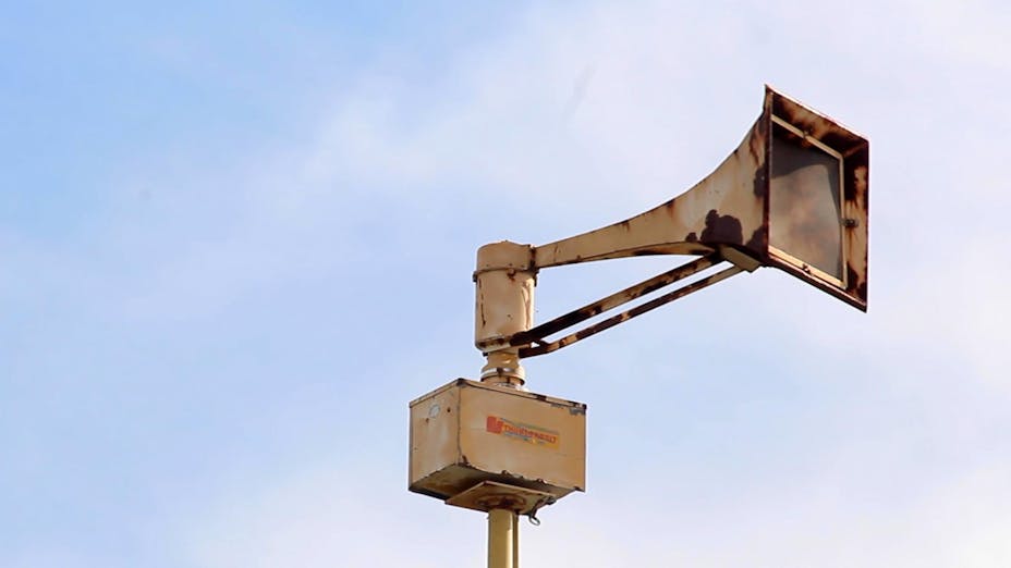 A large air raid siren sits on a tall pole in the sky.