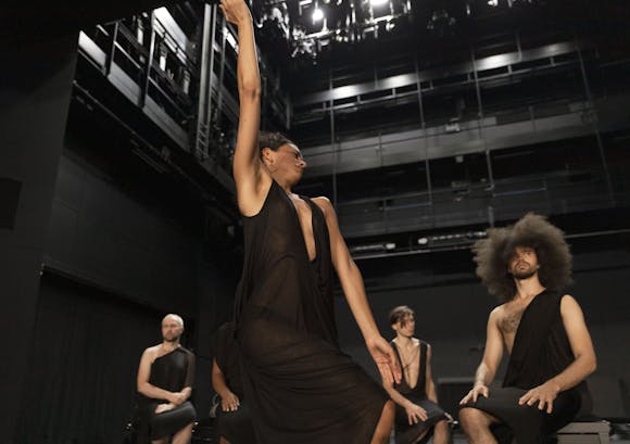A dancer stands on a stage, with their right hand straight over their head. They have light skin and are wearing a black toga. Three other performers of various skin tones and hairstyles sit behind them, on small black benches, also wearing black togas. The upper structure of the stage is visible.