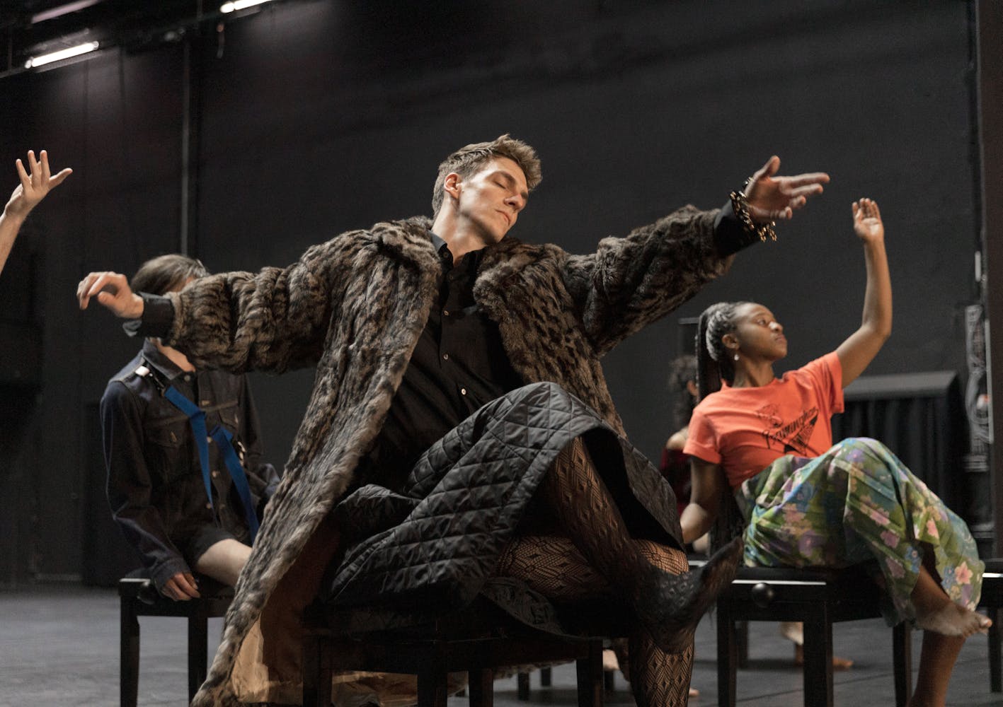 A performer with light skin and short brown hair sits with arms outstretched, wearing a fur coat. Behind them, other performers are also seated with arms outstretched.