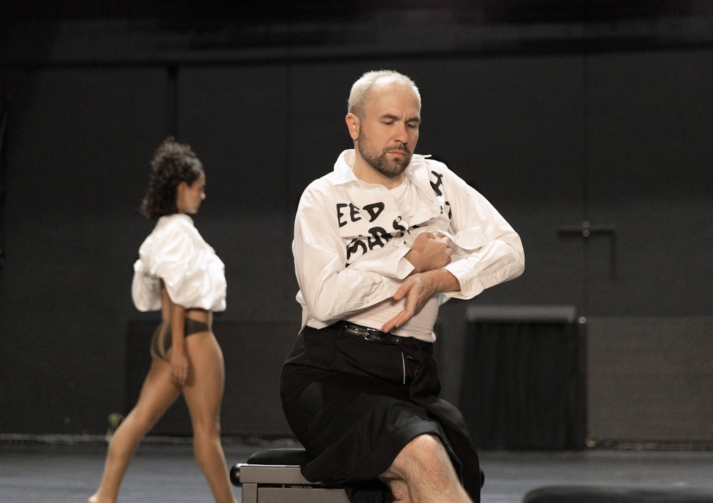 A performer with light skin, short white hair and a short beard sits with their arms folded in front of them, wearing a white shirt with black letters on it. Another performer walks in the background.