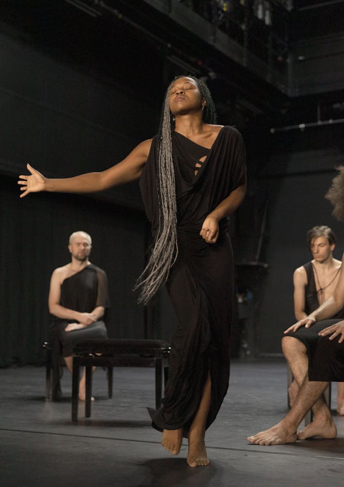 A dancer stands on a stage, with their right hand moving in front of them. They have dark skin, very long braided hair, their eyes are closed, and they are wearing a black toga. Three other performers of various skin tones and hairstyles sit behind them, on small black benches, wearing black togas.