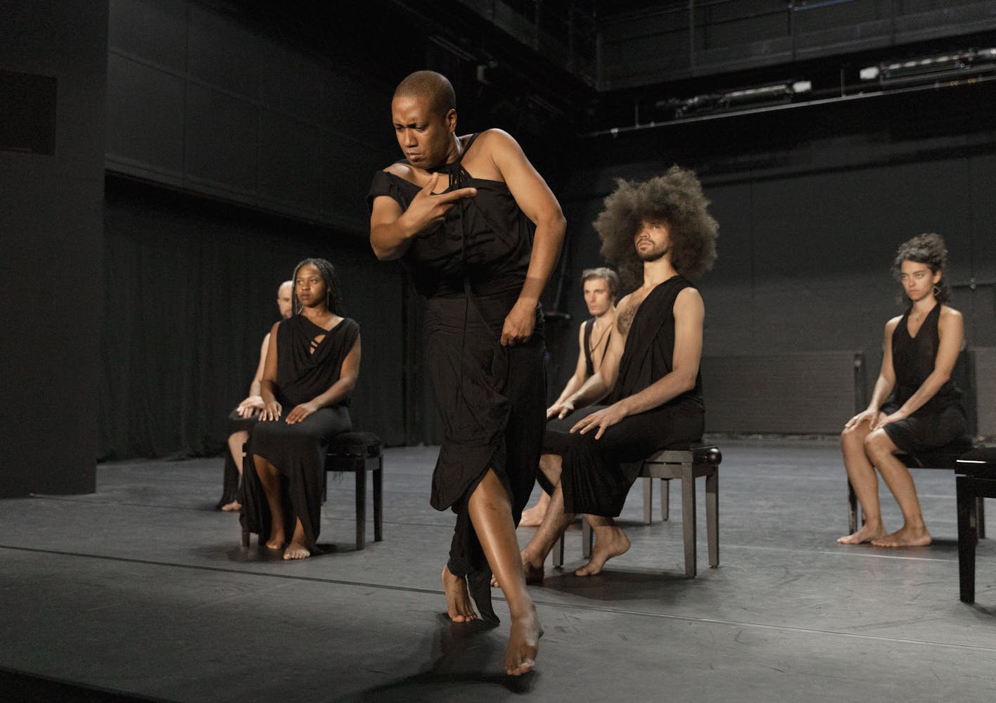 A dancer stands on a stage, with their right hand moving in front of them. They have dark skin, a shaved head and are wearing a black toga. Five other performers of various skin tones and hairstyles sit behind them, on small black benches, wearing black togas as well.