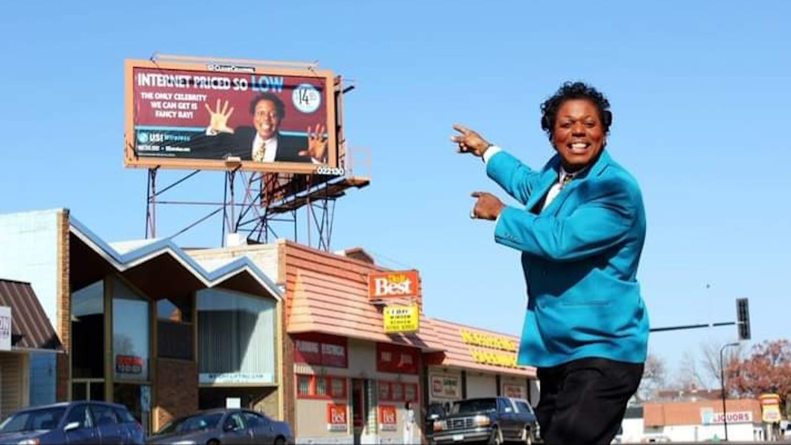A man in a brightly colored suit with a pencil mustach smiles at the viewer while pointing at himself of a billboard behind him.