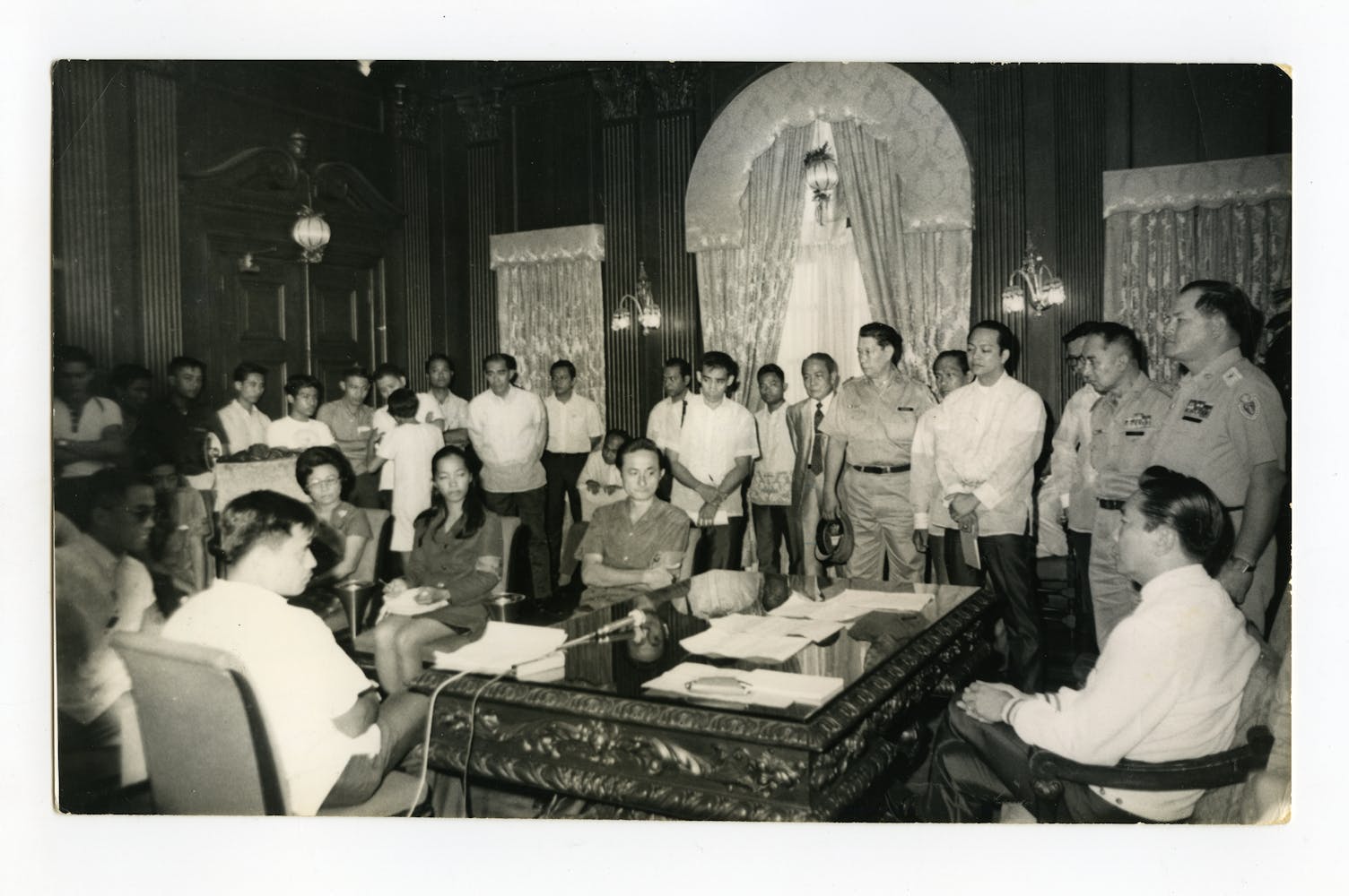 A group of students stand around and sit on a large ornate wooden desk surrounded by men in miliatry uniforms.