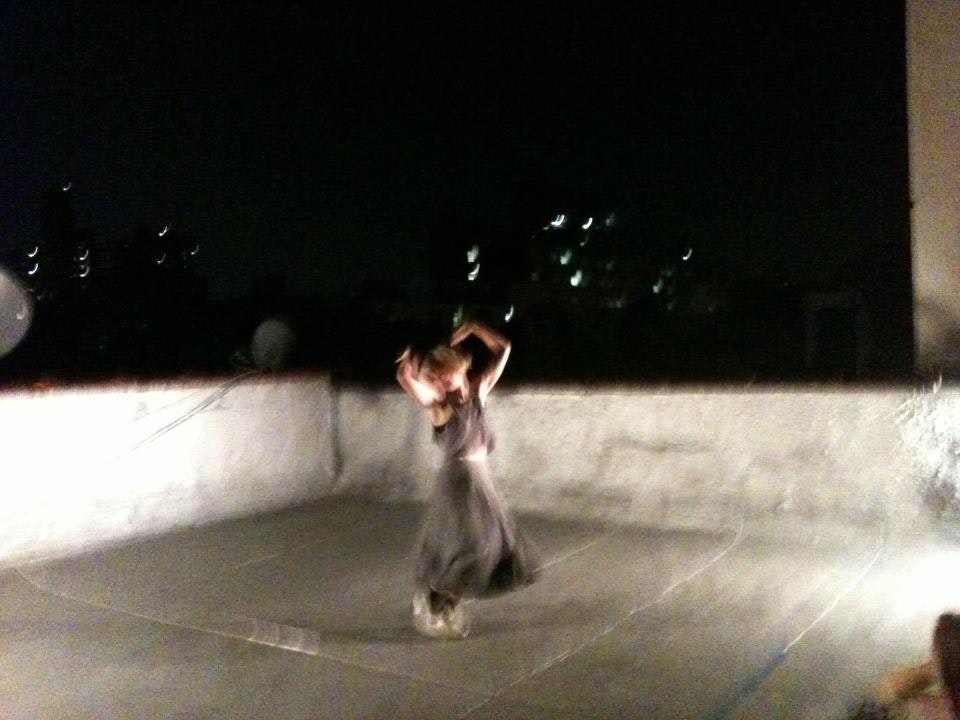 A dancer with white skin and chin-length blond hair dances on a rooftop at night. The image is blurry.