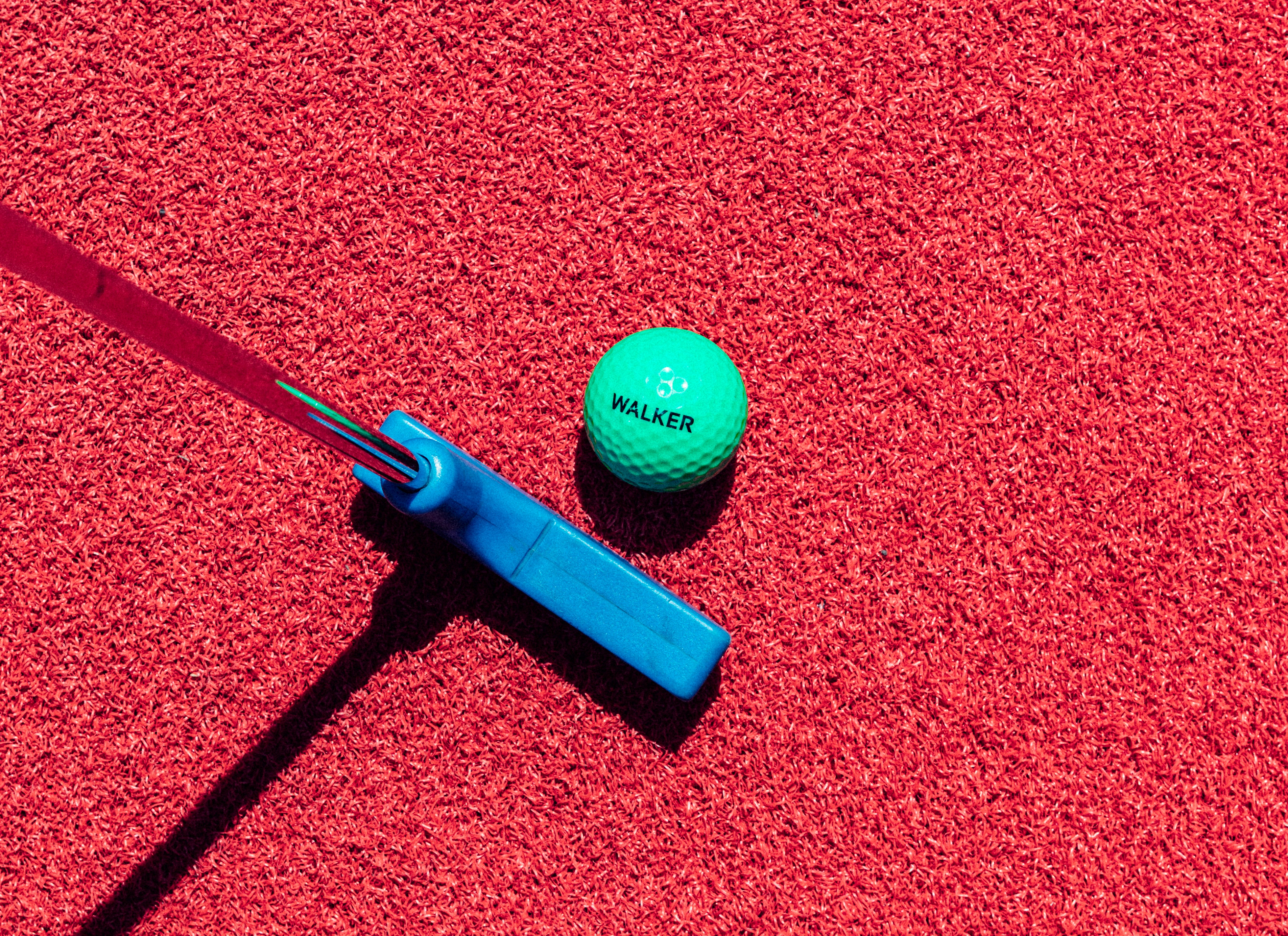 Top view of a green golf ball, blue putting stick red turf.