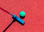 Top view of a green golf ball, blue putting stick red turf.