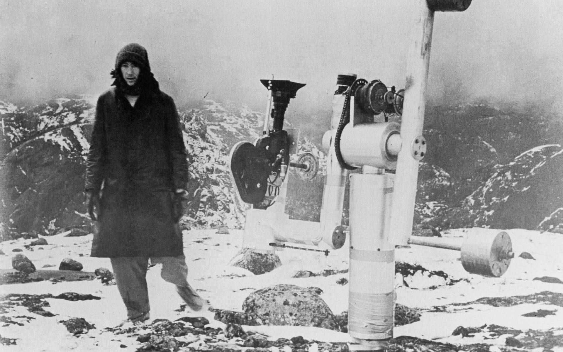 Black and white film still of a man in a foggy snowy mountain top next to a large camera rig