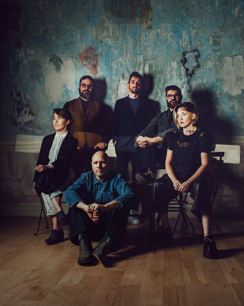 Six performers with light skin pose in front of a textured blue wall.