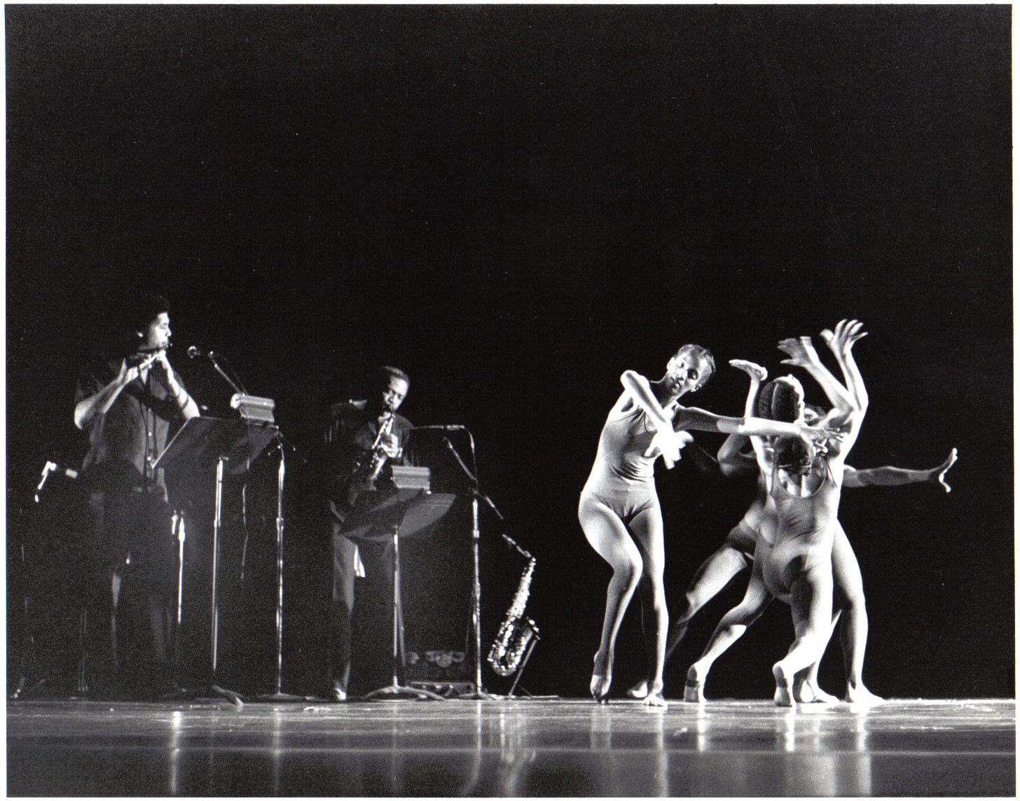 A trio of dancers perform on stage in front of two adults playing a saxiphone and flute.