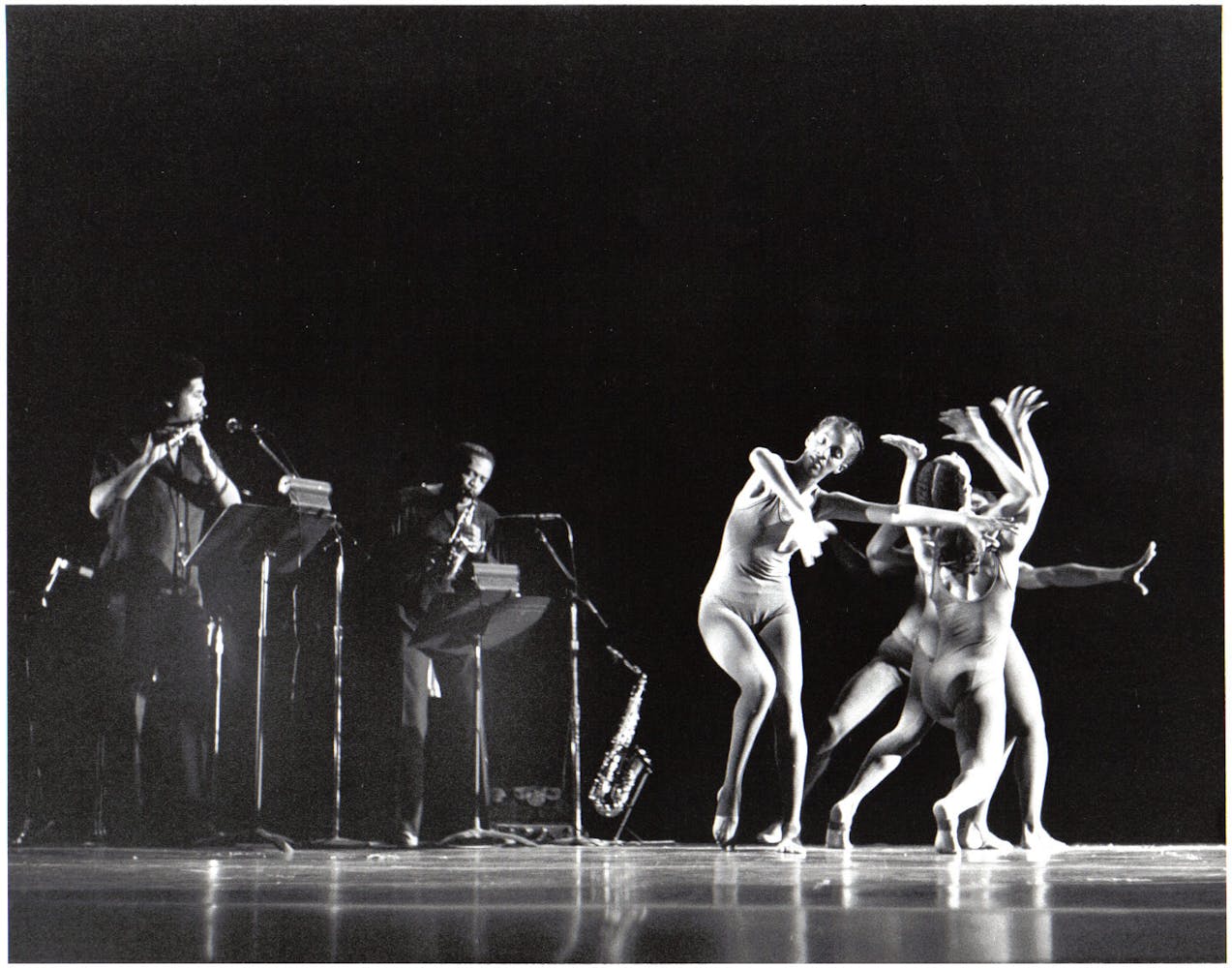 A trio of dancers perform on stage in front of two adults playing a saxiphone and flute.