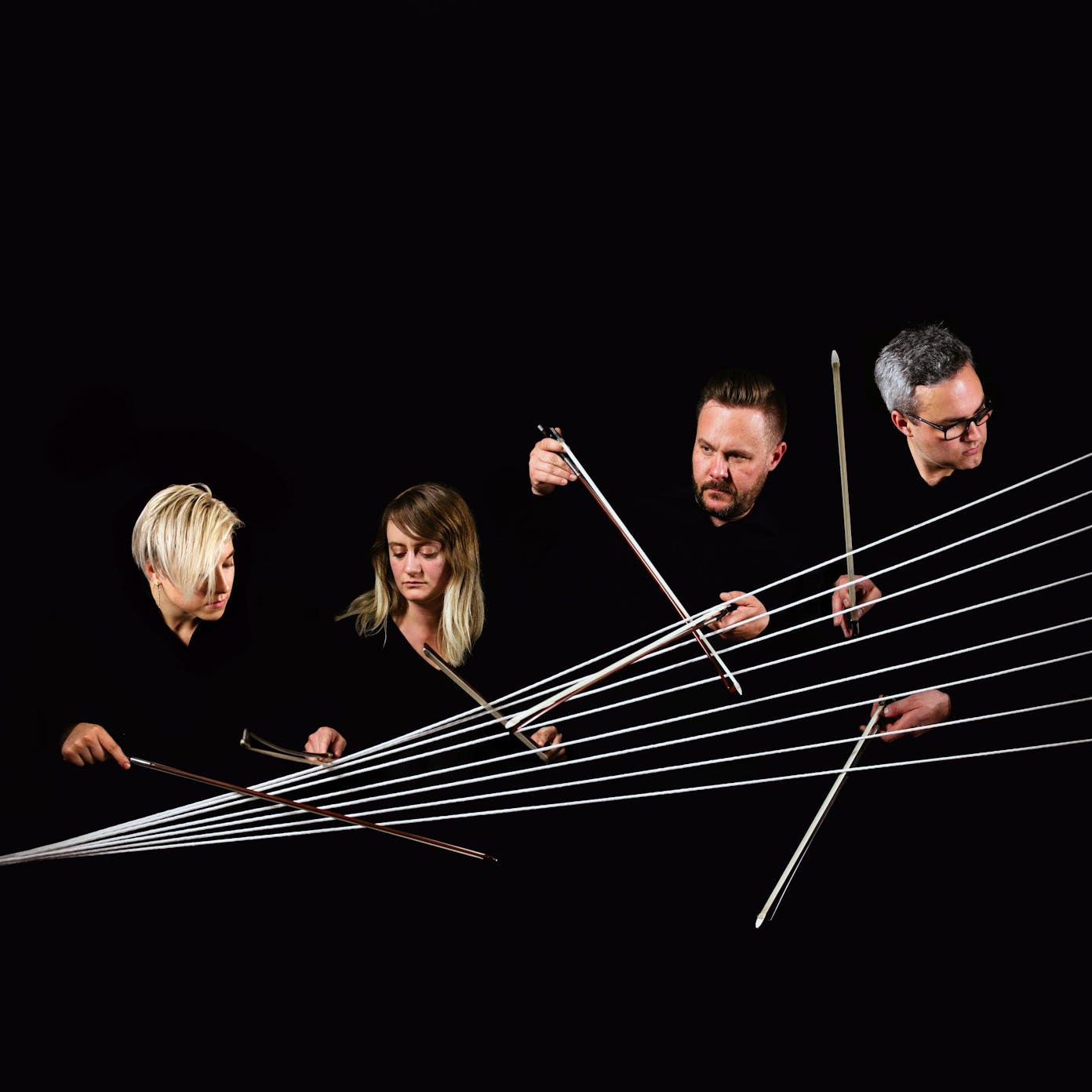 The heads and hands of four people appear out of darkness as they all play a large group of strings.