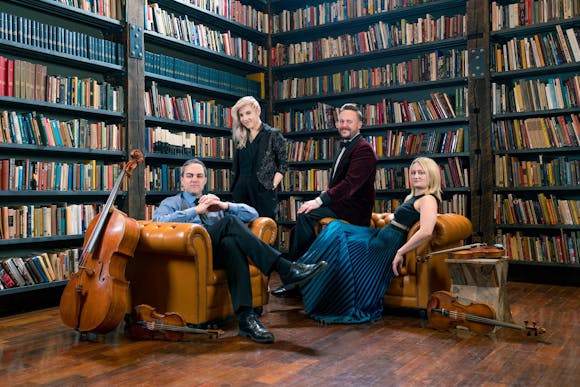 Four people sit next to a cello in a library.