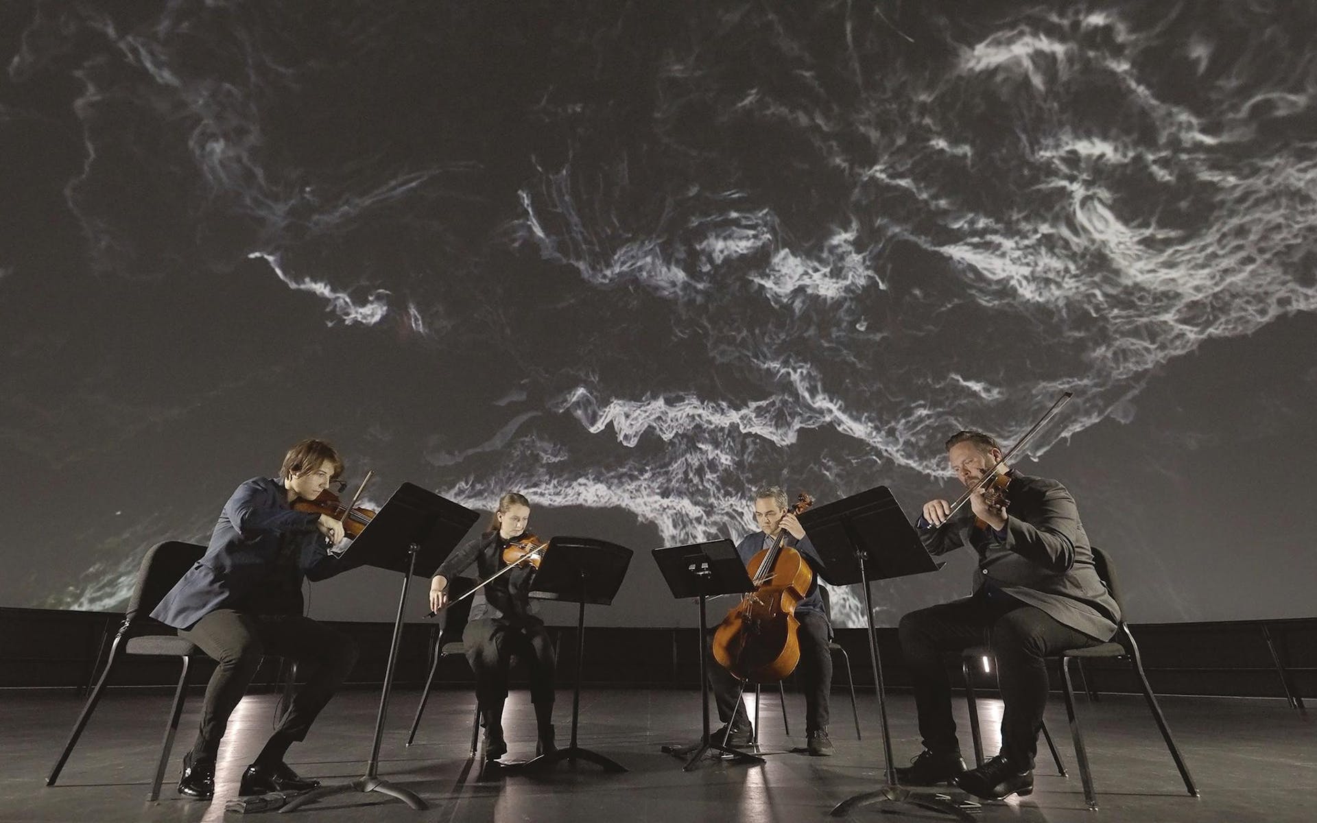 A quartet plays string instraments underneath a large projection of a space rock.