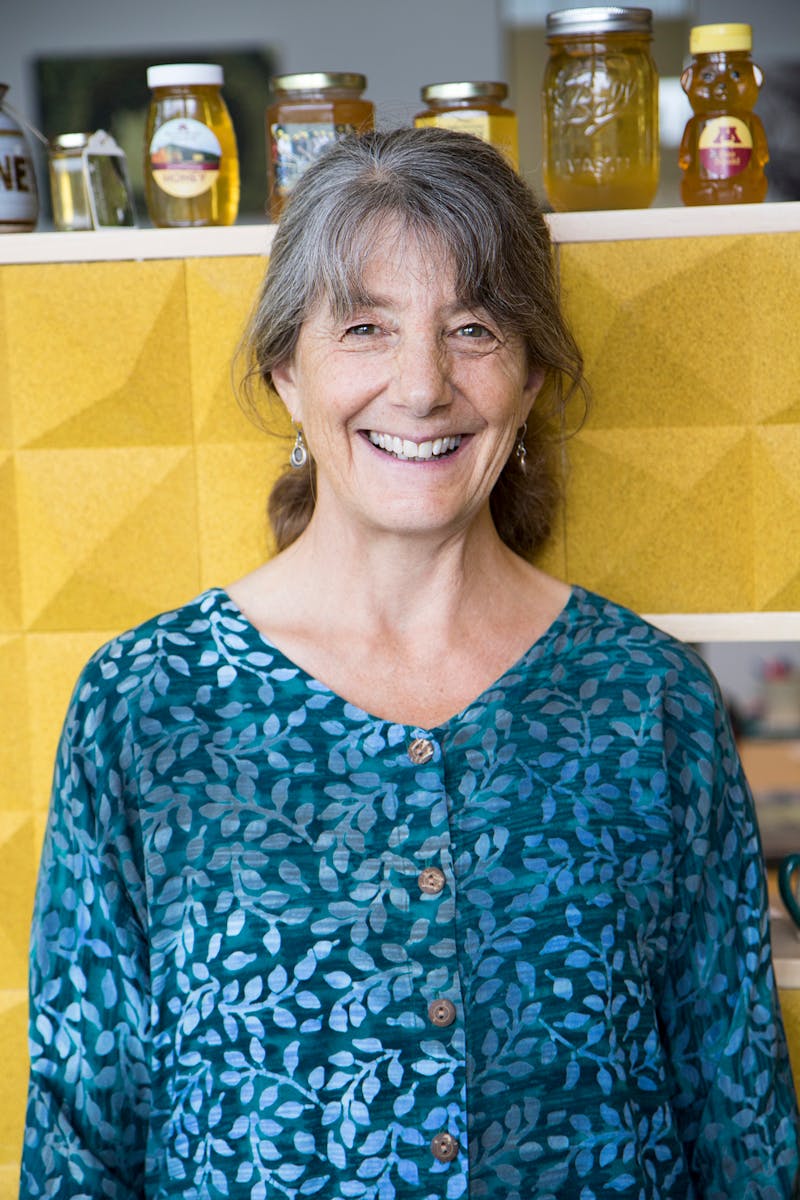 Marla Spivak in a blue blouse standing in front of a yellow wall.