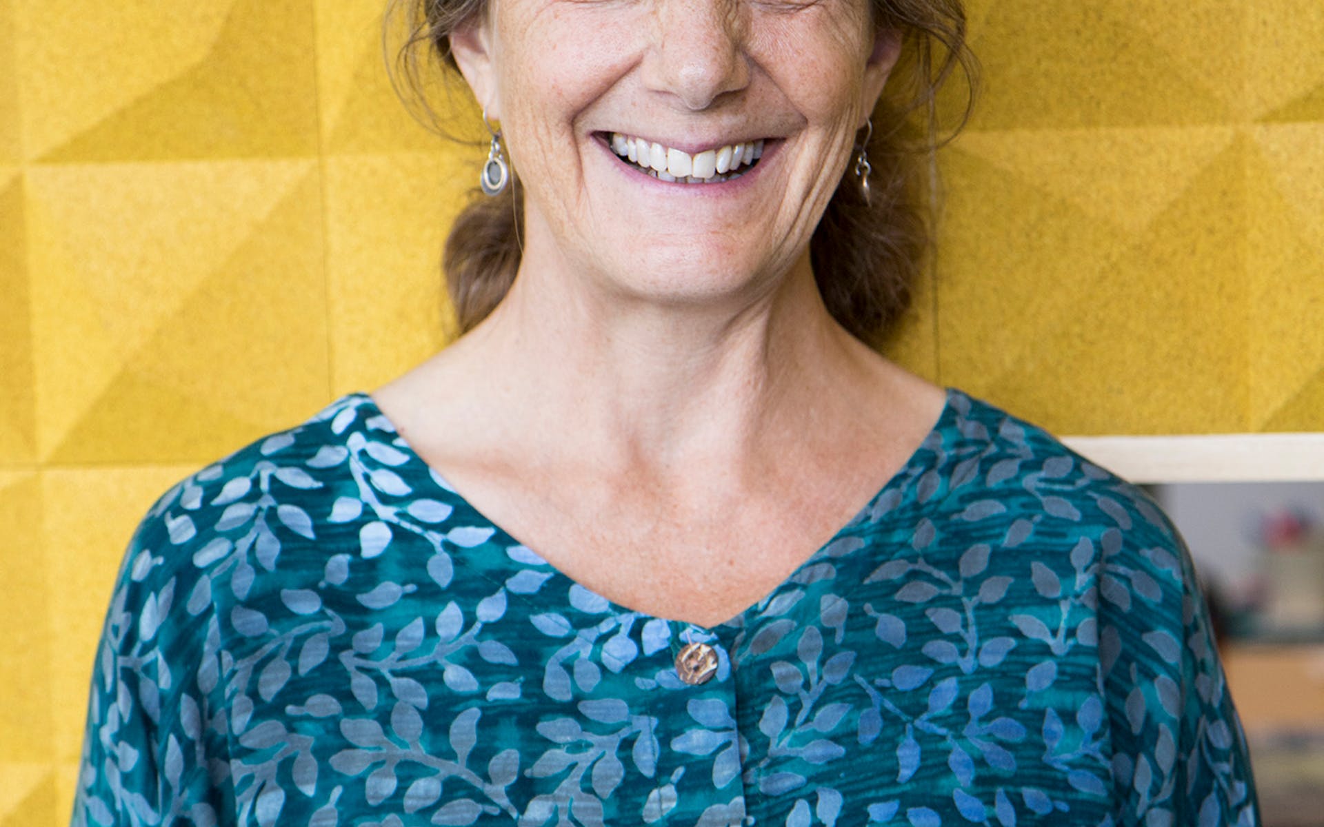 Marla Spivak in a blue blouse standing in front of a yellow wall.
