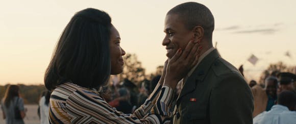 A woman holds the face of a smiling man in a solider's uniform in her hands as she looks into his eyes.