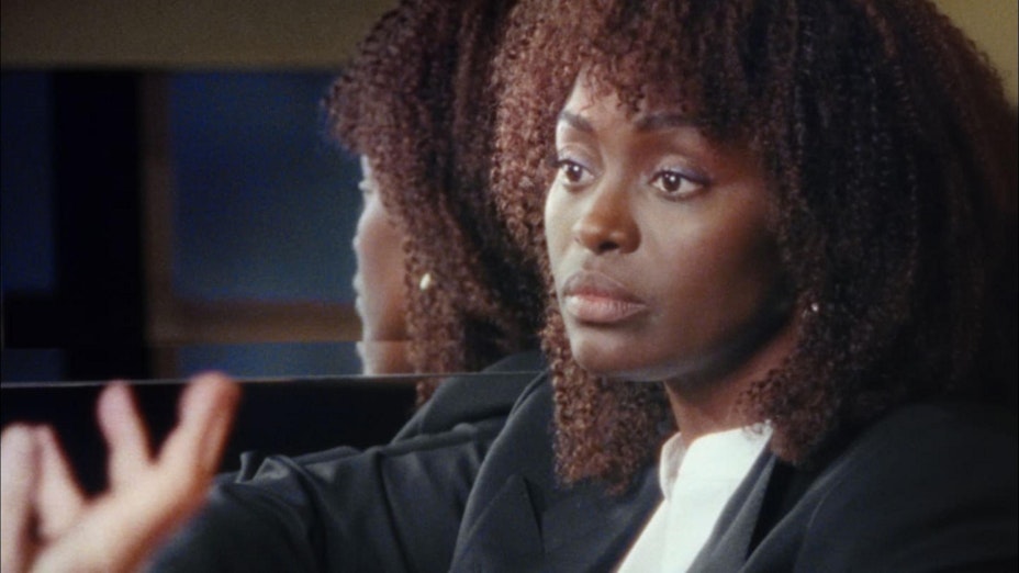 A woman with tight curly hair and bangs wears a black blazer and white shirt. With her back to a mirror, you can also partially see her reflection.