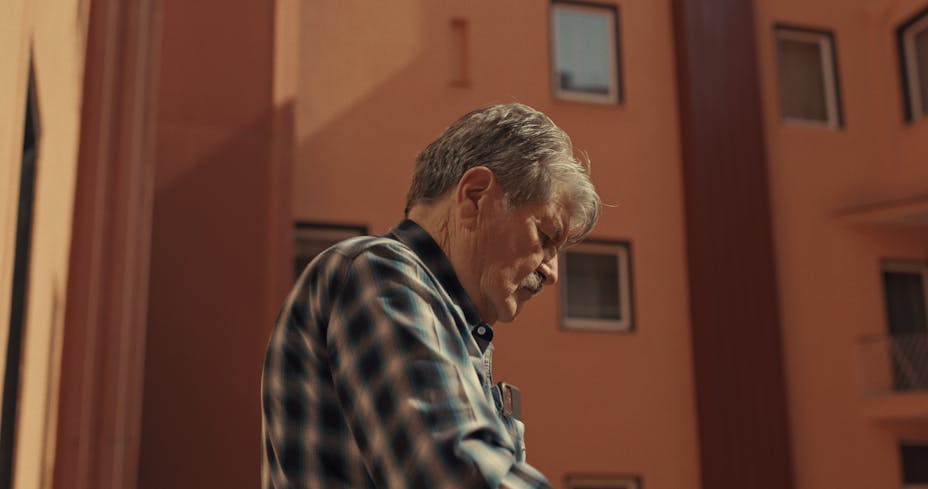 A profile view of light-skinned older man standing in front of a brown building.
